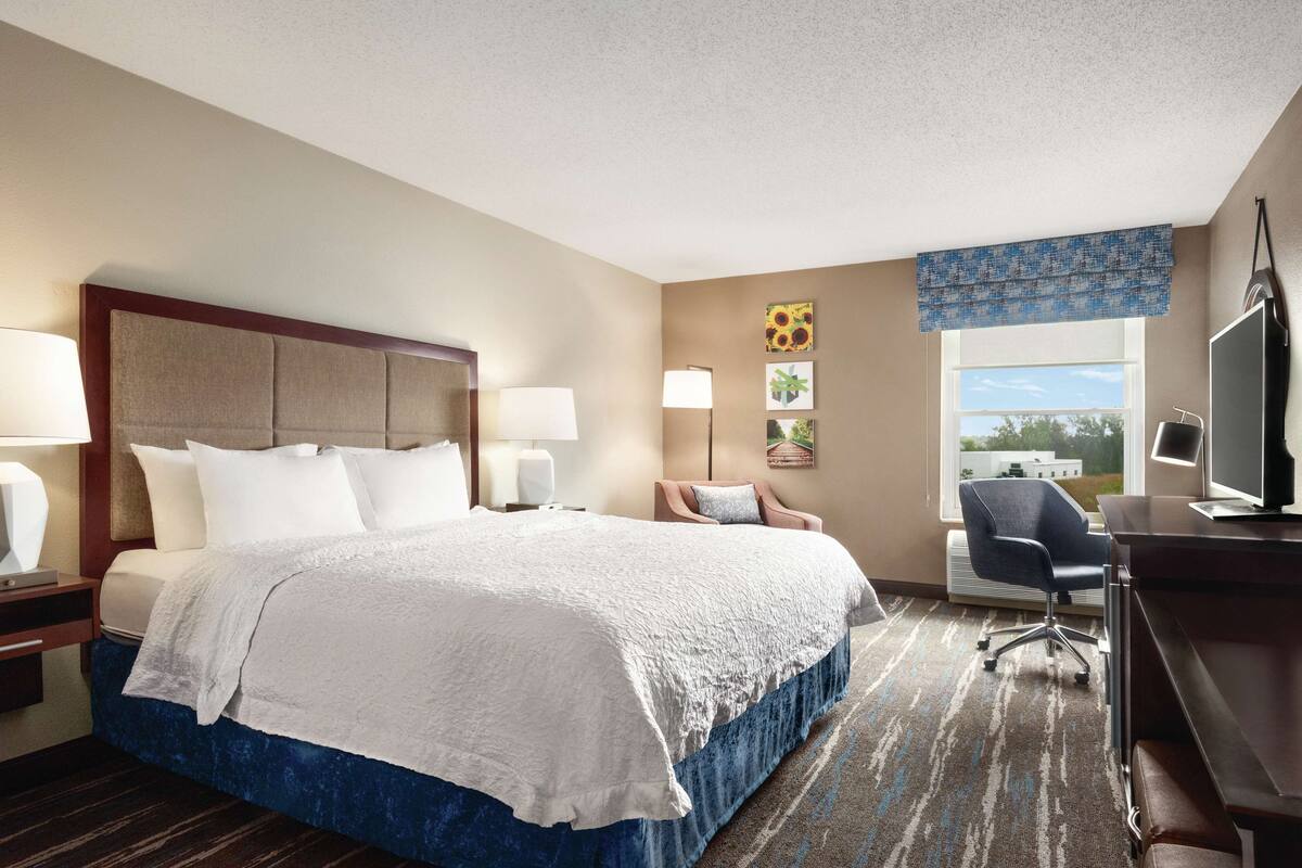 A hotel room with a large bed, two bedside tables with lamps, a desk with a chair, and a large window with a view.