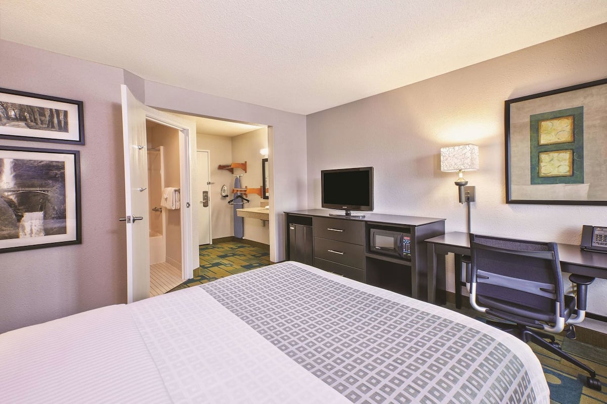 Habitación de hotel con cama, escritorio, silla, televisión y baño visible a través de una puerta abierta.