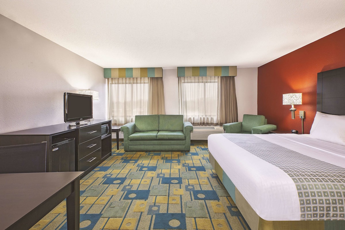 Una habitación de hotel con una cama, un sillón verde, un sofá, un televisor sobre una cómoda y una alfombra estampada.