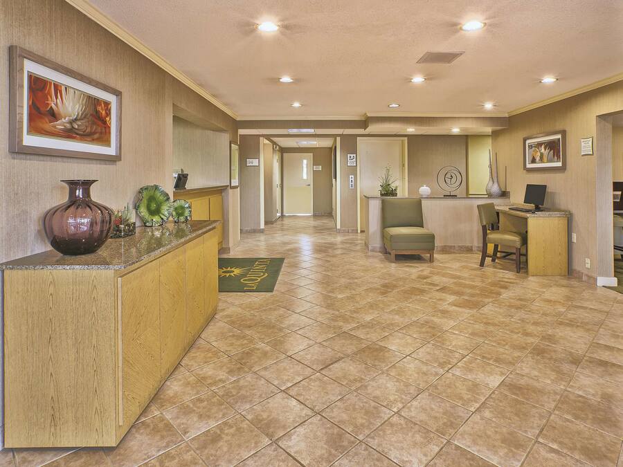 A hotel lobby with a reception desk, a green chair, a wooden reception desk, a vase, and a framed picture on the wall.