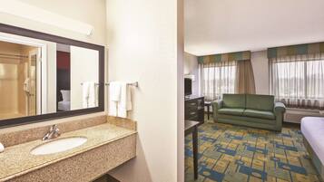 A hotel room with a bathroom featuring a sink and mirror, a green sofa, a bed, and a patterned carpet.