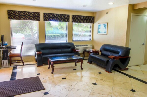 Sala de estar com dois sofás de couro preto, uma mesa de centro de madeira e uma pequena mesinha lateral. Três janelas com persianas, uma porta e um quadro na parede.