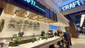A cafe with a modern interior, featuring a counter with plants, a menu display, and a ceiling with recessed lighting.