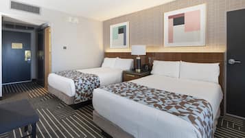 A hotel room with two beds, a desk, and a chair. There are two bedside tables with lamps, a wall-mounted phone, and a door with a handle.