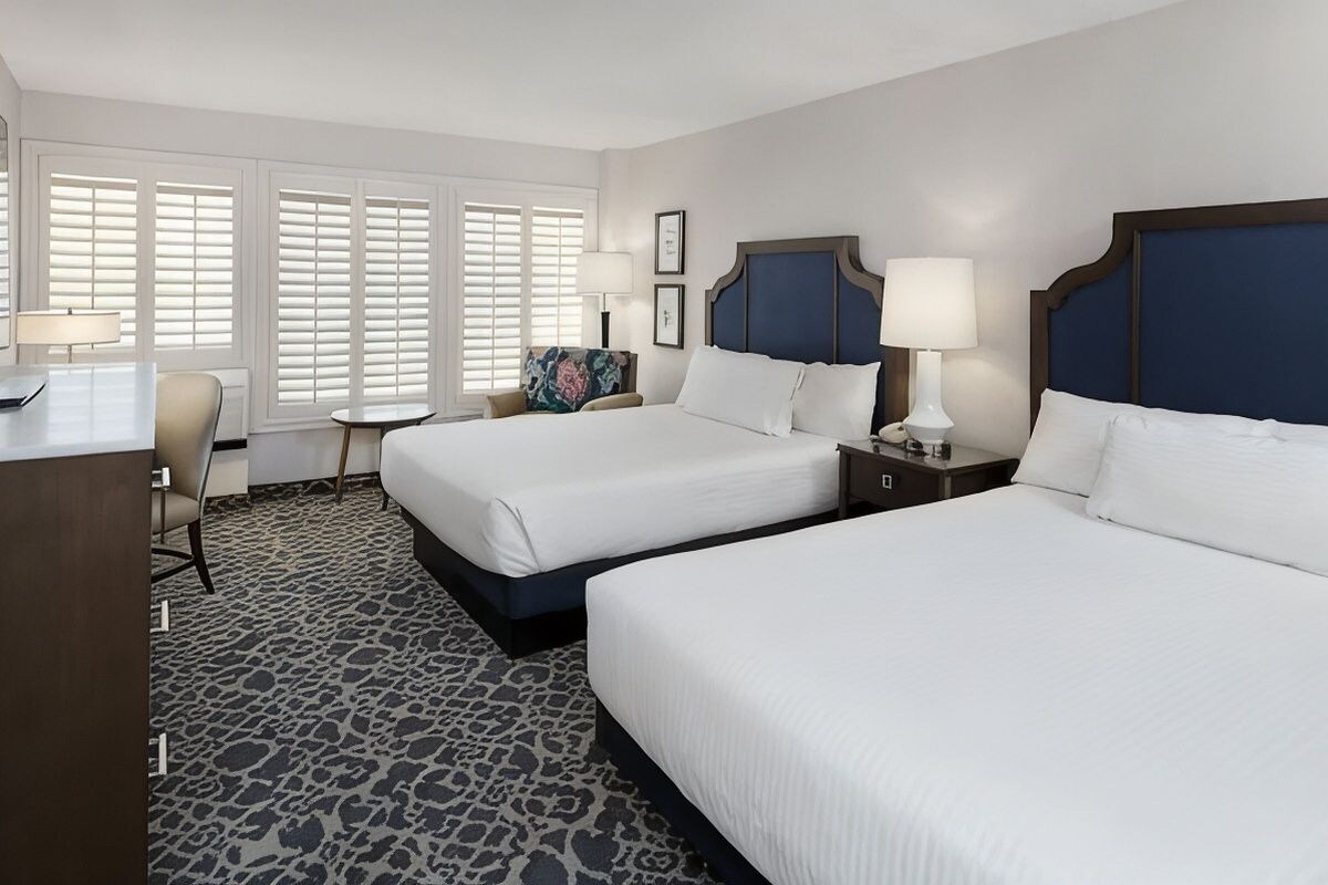 A hotel room with two beds, a desk, a chair, and a small table.