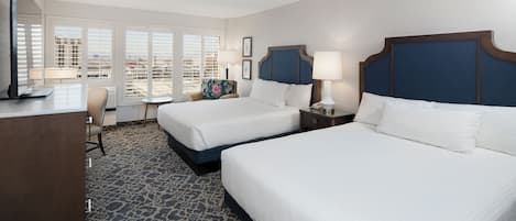 A hotel room with two beds, a desk, a chair, and a large window with blinds.
