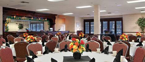 Un salón de conferencias con mesas dispuestas para una reunión, un escenario con un cartel y una zona de recepción.