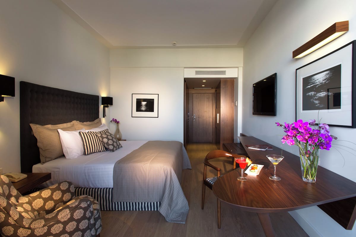 A hotel room with a large bed, a desk with two chairs, a small table with drinks, and a vase of flowers.
