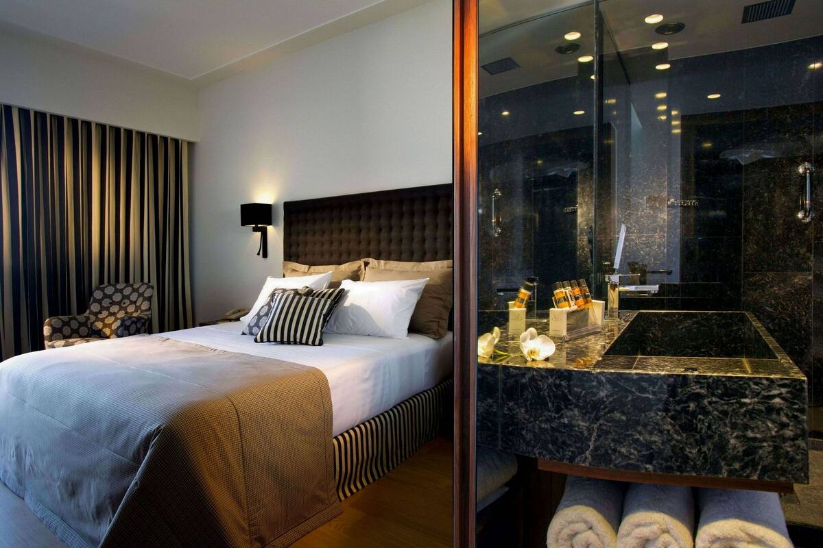 A modern hotel room with a large bed, a headboard, a nightstand with toiletries, a chair, and a bathroom with a marble sink and mirror.