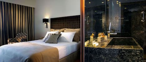 Una habitación de hotel moderna con una cama grande, cabecera, mesita de noche con artículos de tocador, una silla y un baño con lavamanos y espejo de mármol.