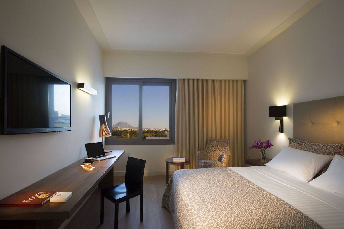 A hotel room with a bed, a desk with a laptop, a chair, a TV mounted on the wall, and a window with a view of a mountain.