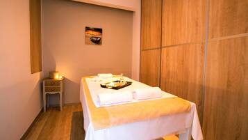 A spa room with a massage table, towels, a tray with amenities, a small bedside table with a lamp, and a painting on the wall.