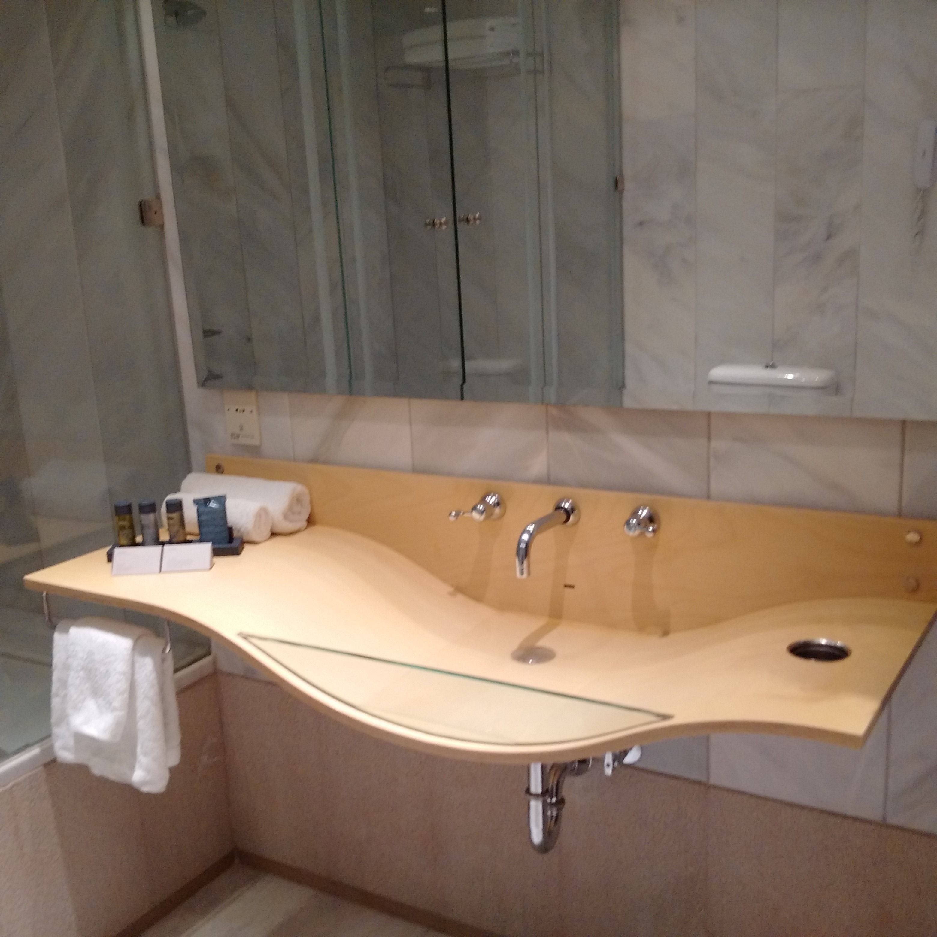 Un baño con un lavamanos curvo, una cabina de ducha con mampara de vidrio y un revestimiento de mármol en la pared.