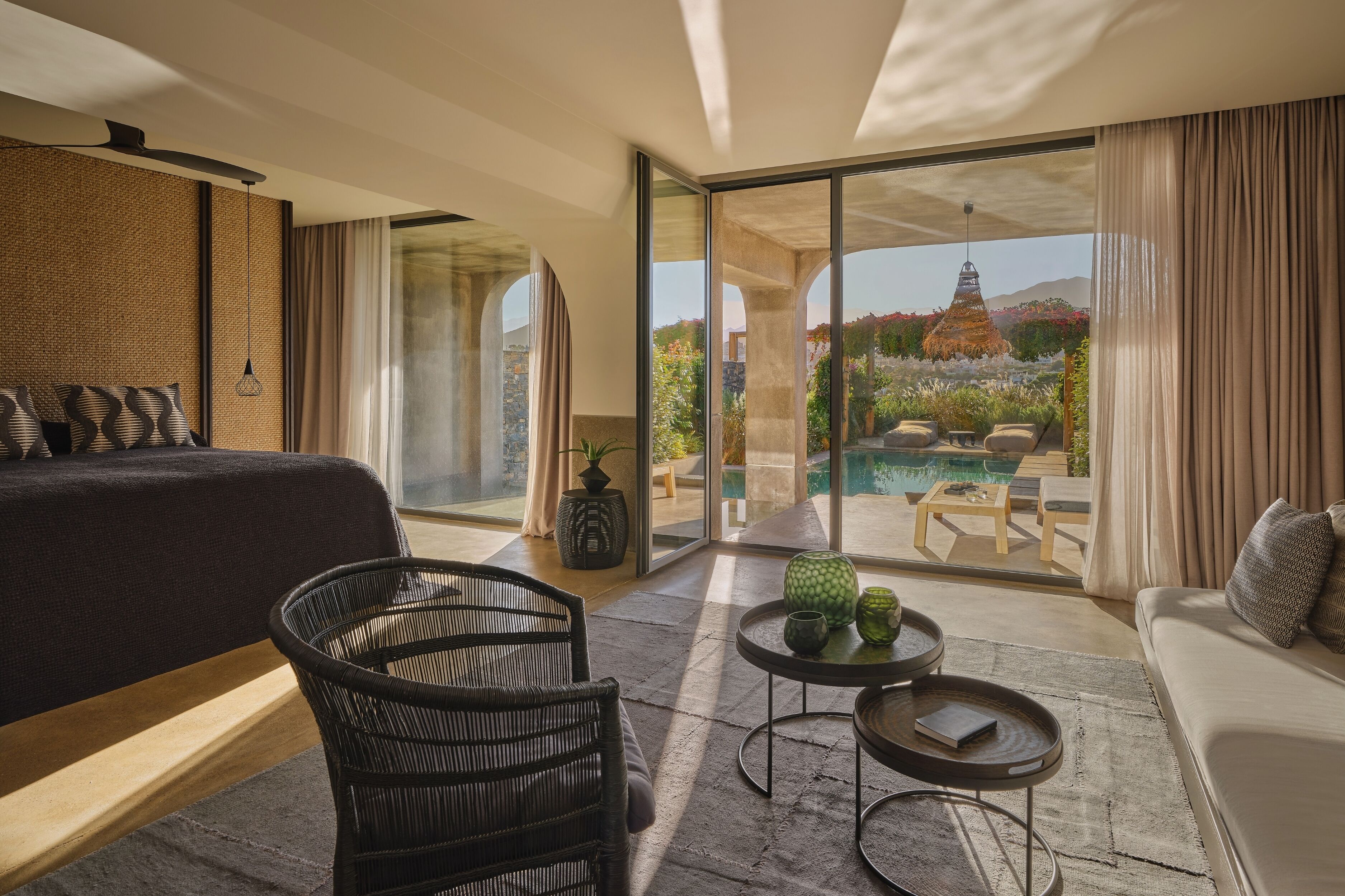 Habitación de hotel moderna con una cama grande, zona de estar con sofá y sillones, una mesita con objetos decorativos, y vistas a una piscina y jardín a través de amplios ventanales.