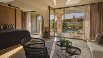 Habitación de hotel moderna con una cama grande, zona de estar con sofá y sillones, una mesita con objetos decorativos, y vistas a una piscina y jardín a través de amplios ventanales.