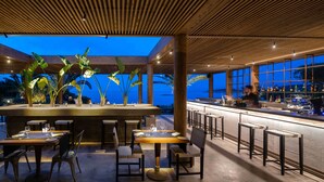Een buitenterras met tafels en stoelen, een bar en uitzicht op de oceaan in de schemering.