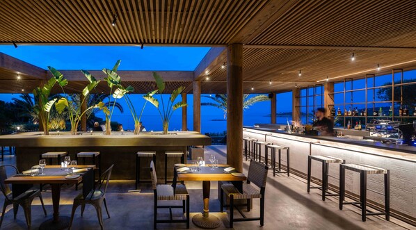 An outdoor dining area with tables and chairs, a bar, and a view of the ocean at dusk.
