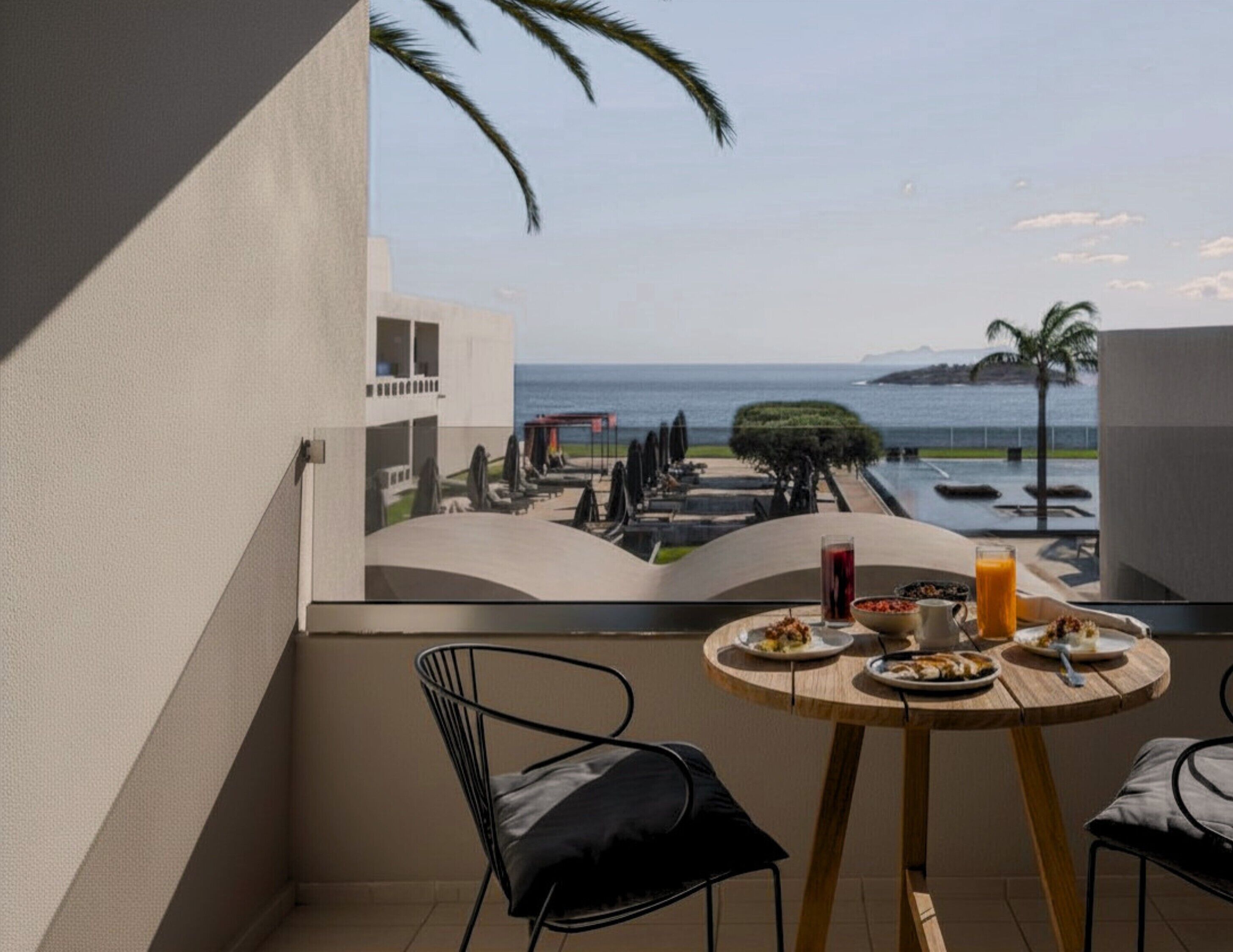 A balcony with a table set for breakfast, overlooking a pool and a view of the sea.