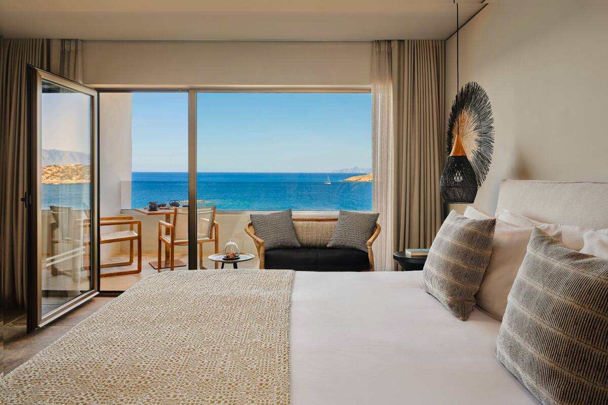 Hotelkamer met een groot bed, uitzicht op zee, een balkon met een tafel en stoelen, en een decoratief kunstwerk aan de muur.