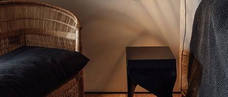 Una silla de mimbre con un cojín oscuro, una mesita negra y una pared con una grieta visible.