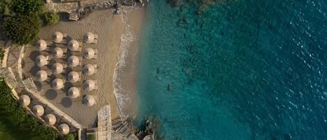 Vista aérea de una playa con agua turquesa cristalina, un camino de piedra que conduce a una hilera de sombrillas y afloramientos rocosos.