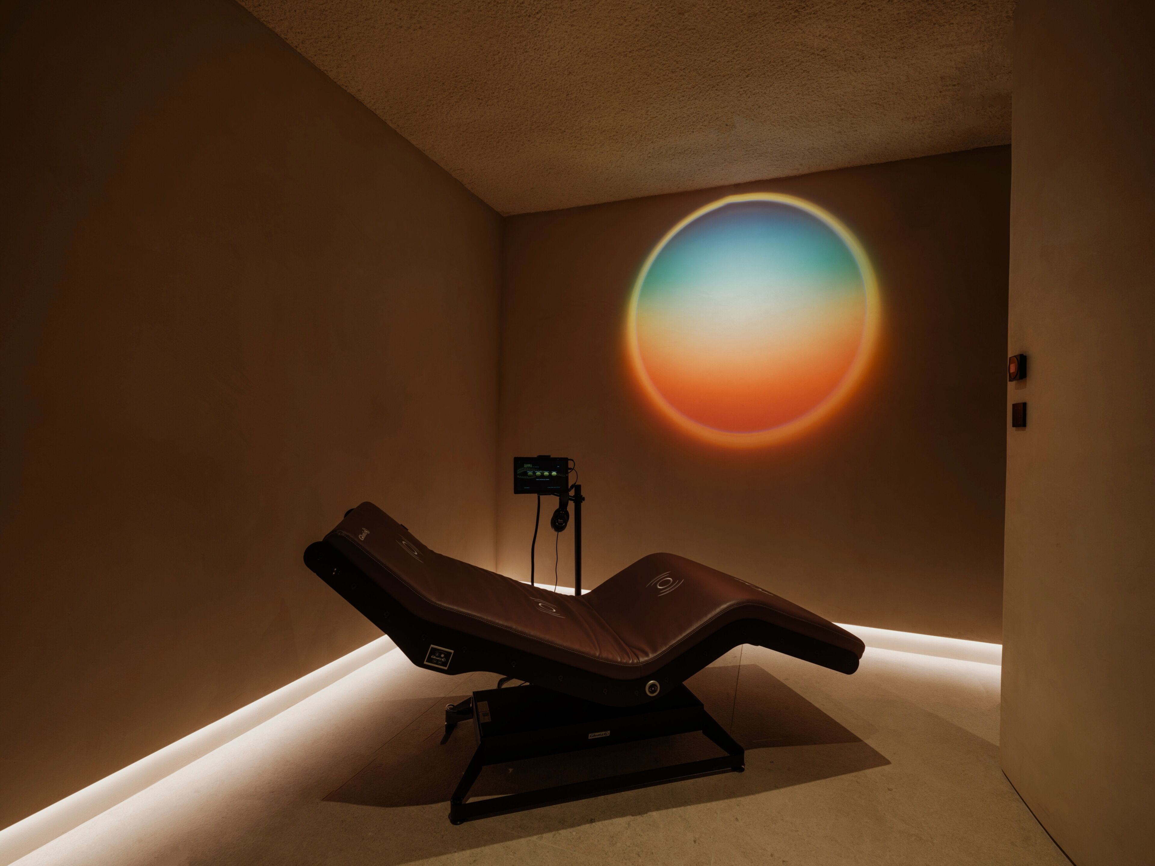 Un cuarto de terapia de masajes con una sola camilla reclinable, un equipo de masaje fijado a la pared y una gran proyección de luz circular.