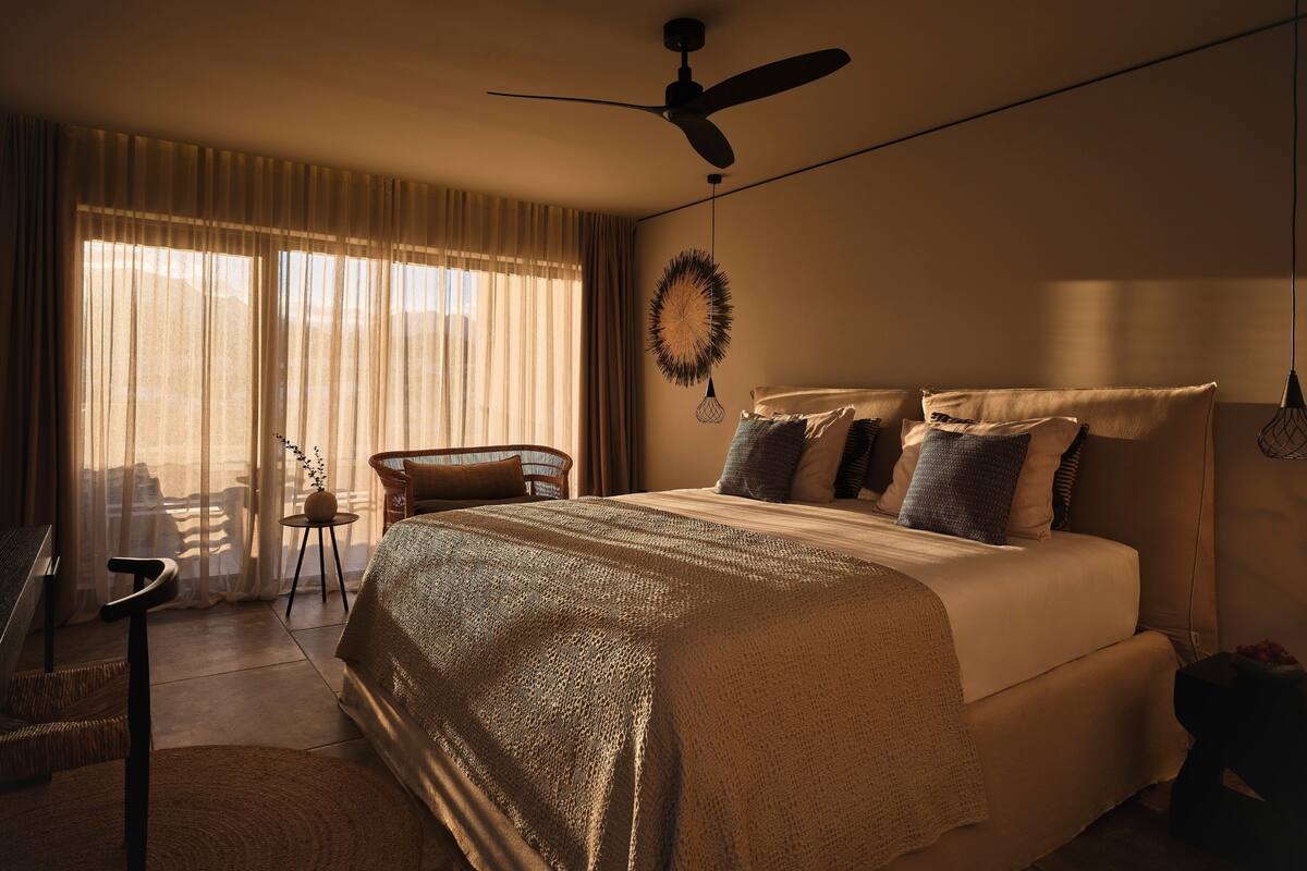 Een slaapkamer met een groot bed, een plafondventilator, uitzicht op het landschap door dunne gordijnen en een klein, rond tafeltje met een stoel.
