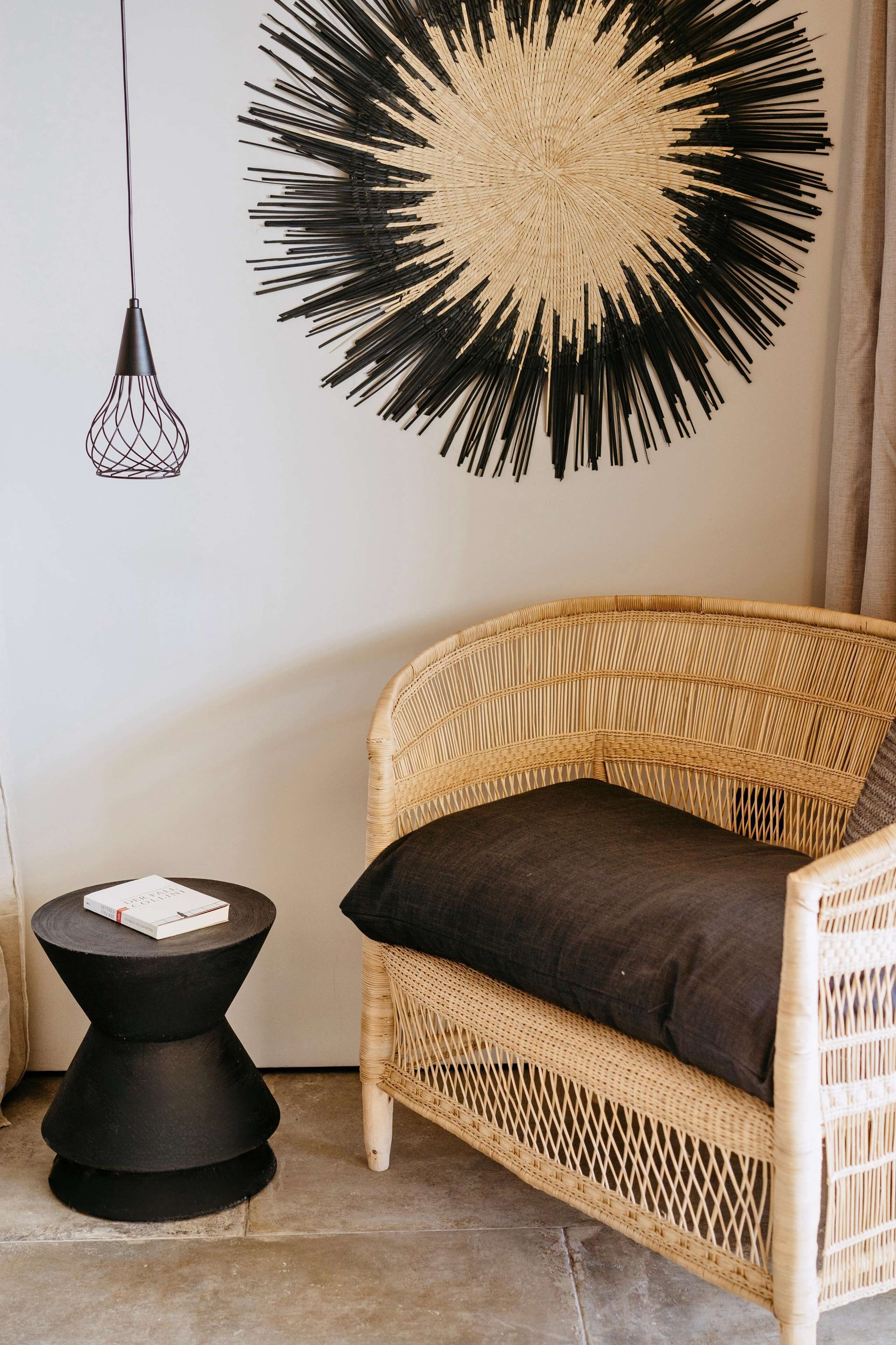 A wicker chair with a dark cushion, a black side table with a book, and a wall art piece.