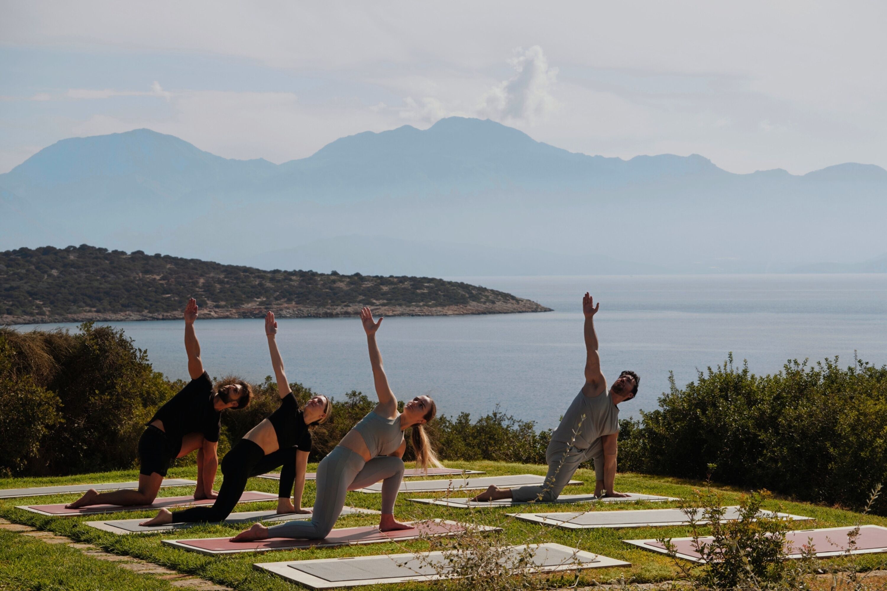 A group of people practicing yoga on mats outdoors with a mountainous landscape in the background.