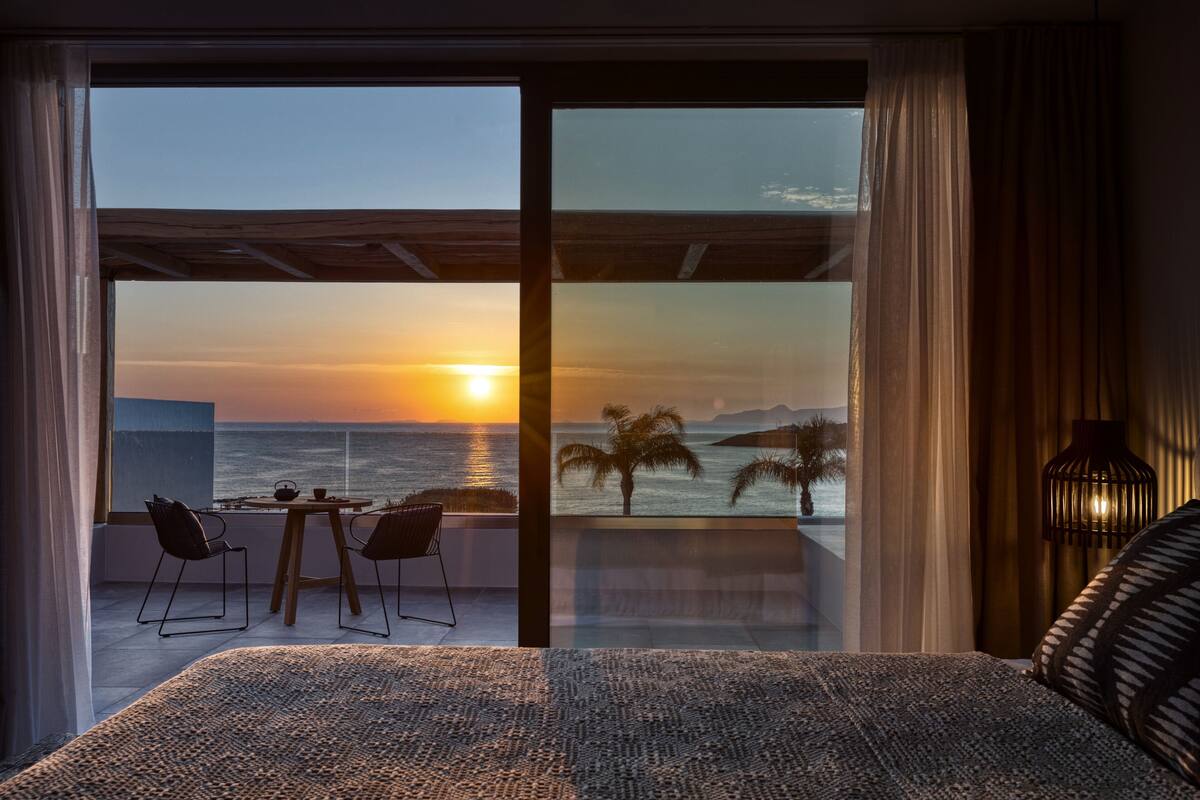 Een slaapkamer met een grote schuifglazen deur die leidt naar een balkon met uitzicht op de ondergaande zon boven de oceaan.