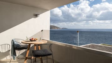 Un balcon avec une table ronde en bois, deux chaises en métal noir et un saladier rempli de fruits. Le balcon donne sur une étendue d’eau et sur des collines lointaines.