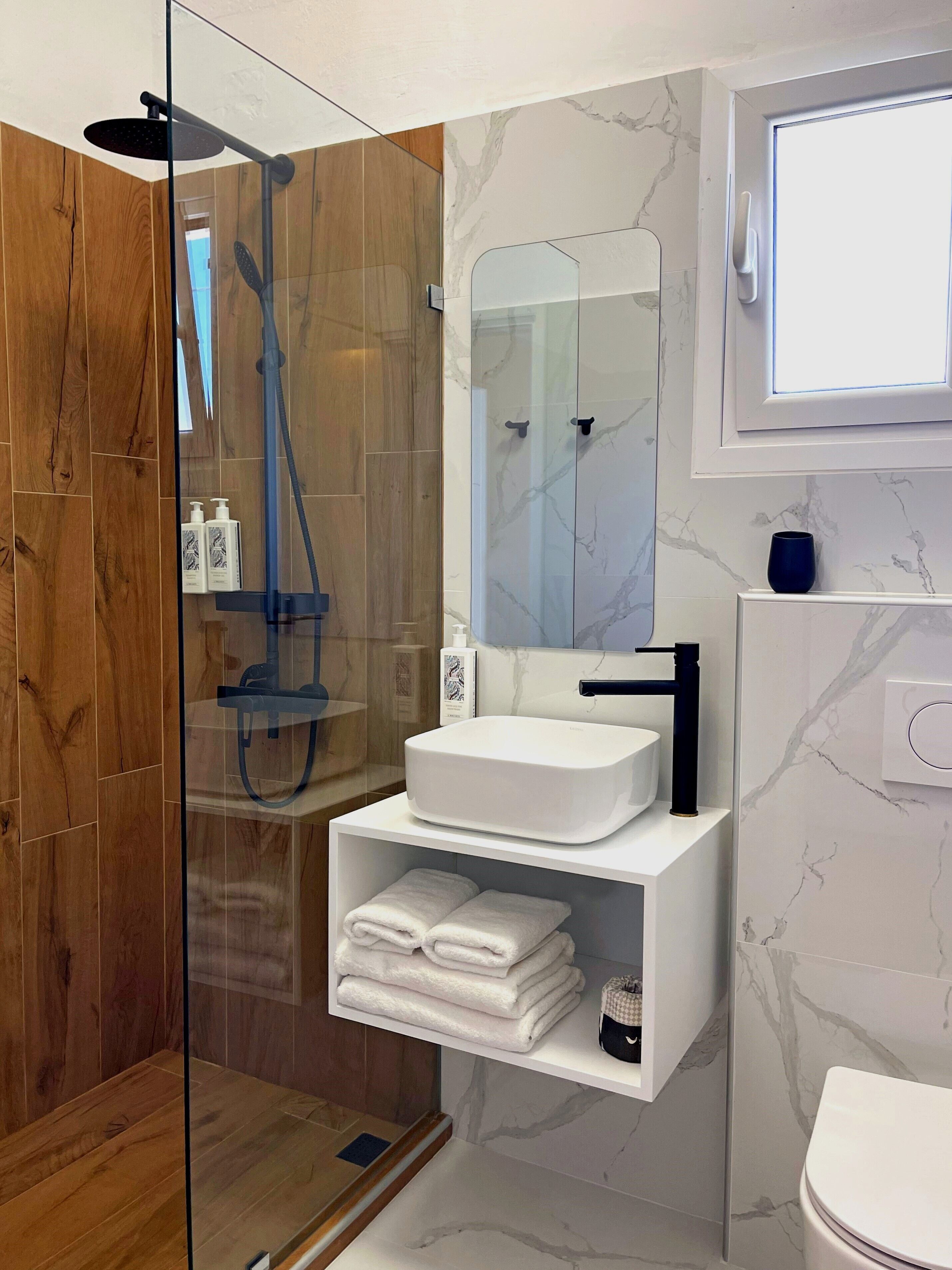 Une salle de bain moderne avec une douche à parois de verre, un lavabo blanc doté d’un robinet noir et des murs à motifs de marbre.