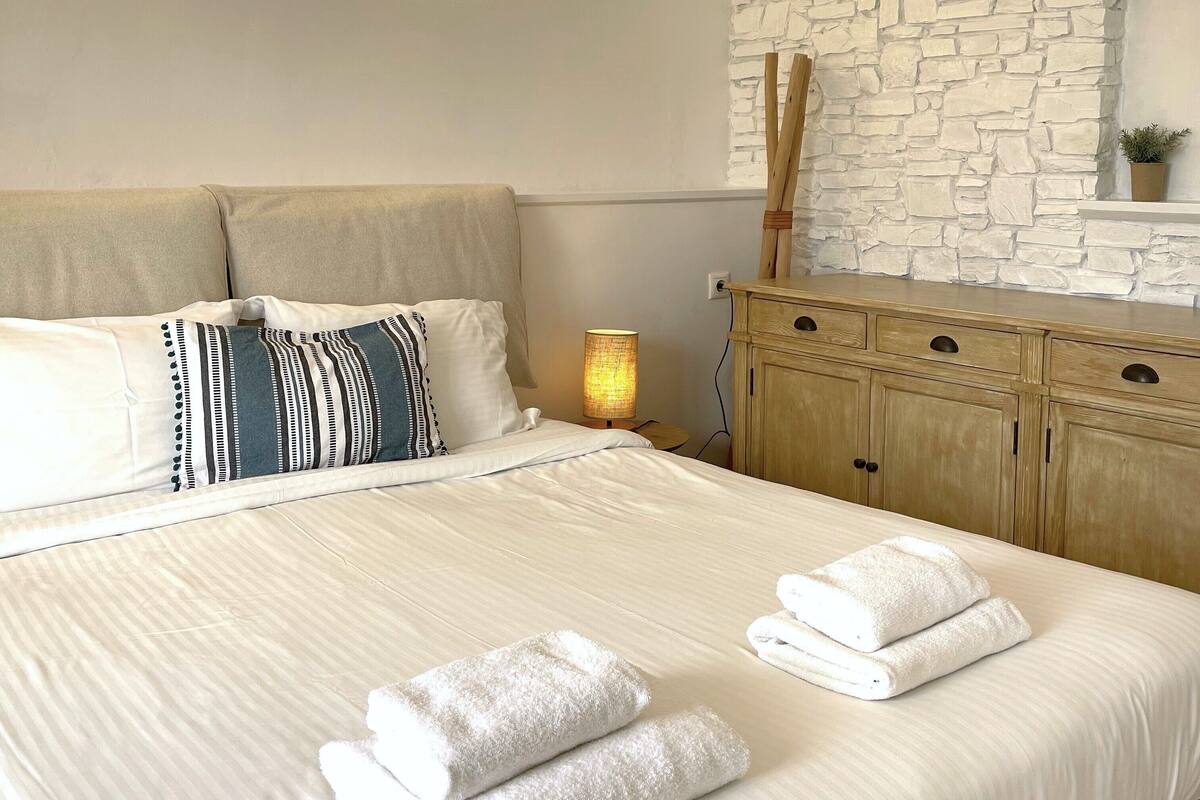 Una cama bien hecha con sábanas blancas y dos almohadas, un aparador de madera con un espejo, y una pared de piedra con una pequeña planta.
