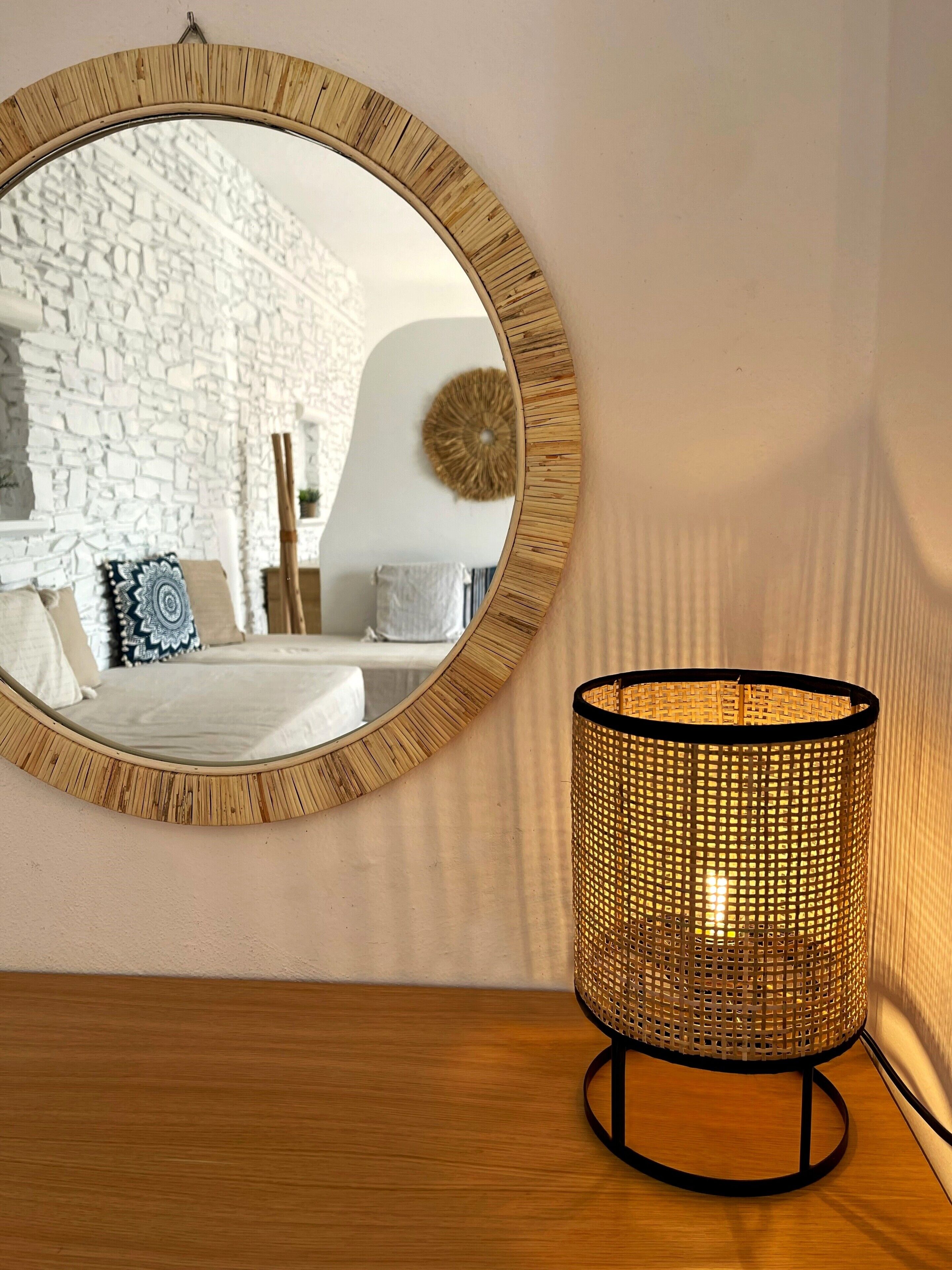 A round mirror with a wooden frame, a woven lamp, and a stone wall in the background.