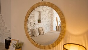 A bedroom with a stone wall, a round mirror, a bed, and a bedside lamp. - Adonis Hotel & Apartments (Paros)