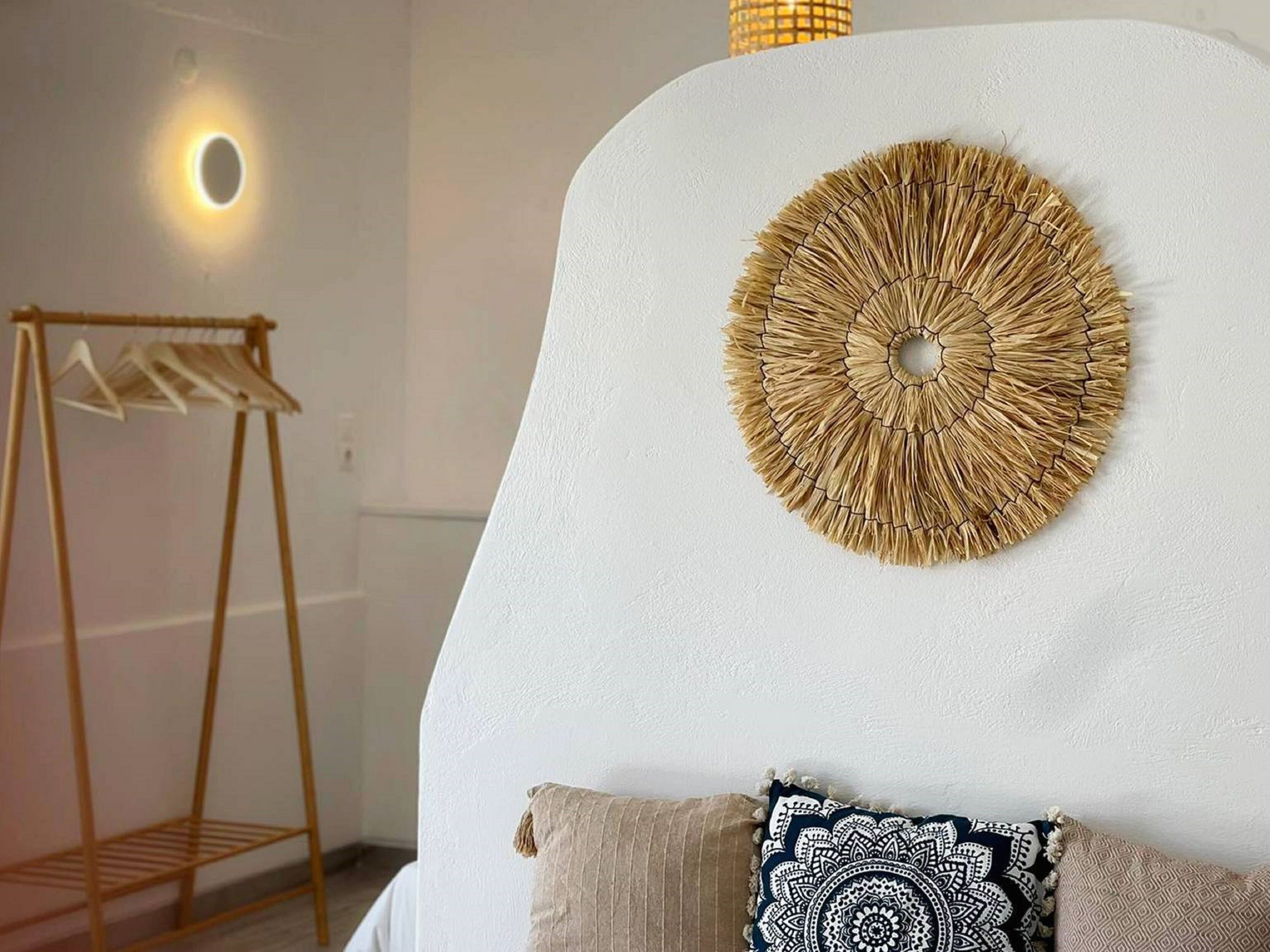 A room with a white wall, a circular woven wall art piece, a wooden clothes rack, and a patterned pillow.