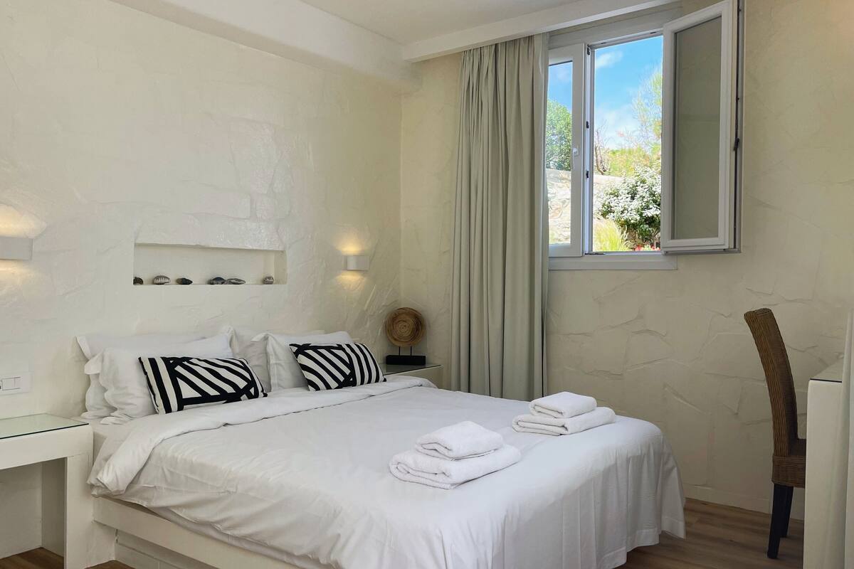 Un dormitorio con una cama, ropa de cama blanca, una mesita de noche, una lámpara de pared, una ventana con cortinas y piso de madera.