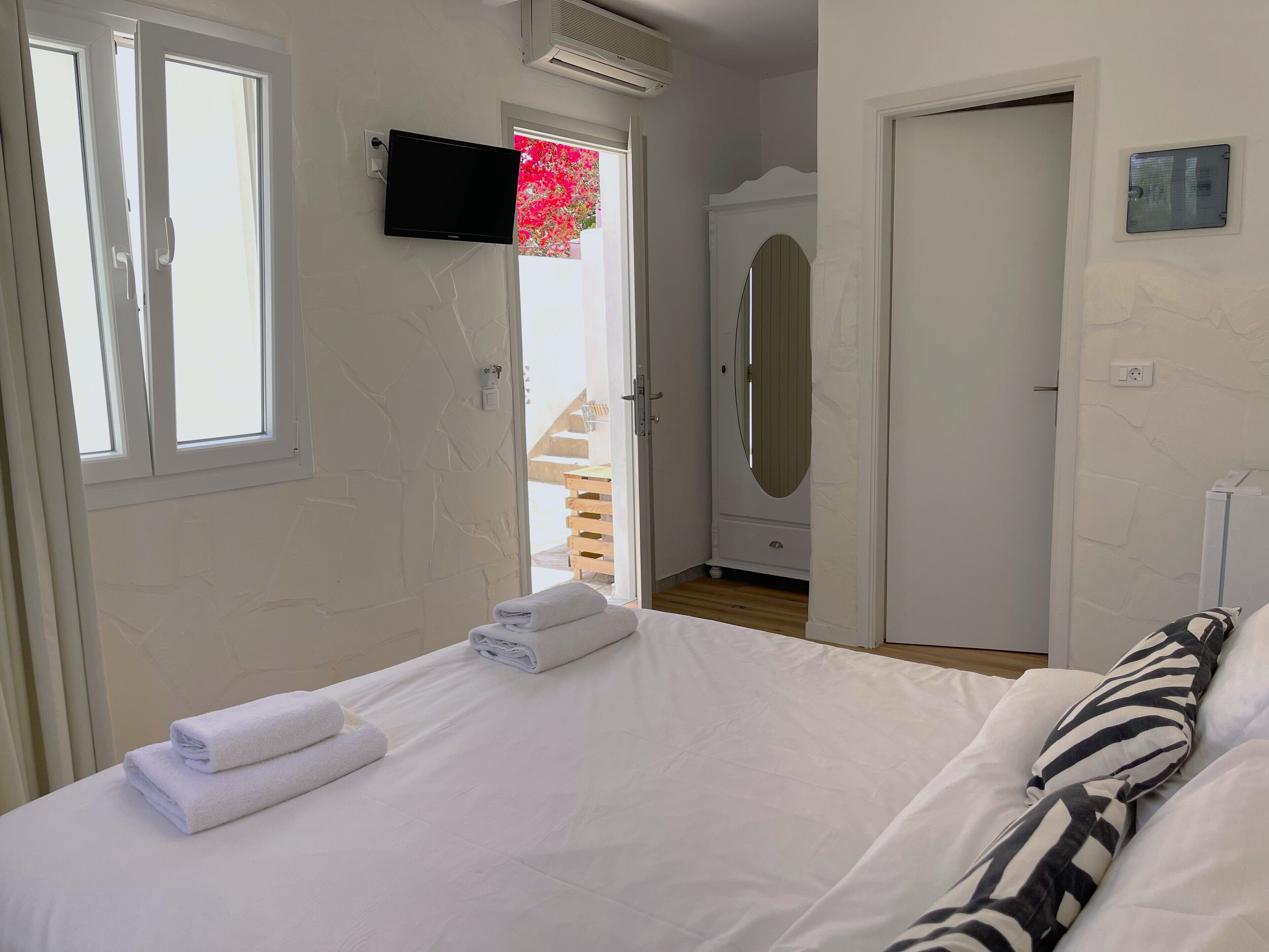 Ett hotellrum med en säng, en tv som är monterad på väggen och en dörr som leder ut till en balkong med utsikt över blommor.