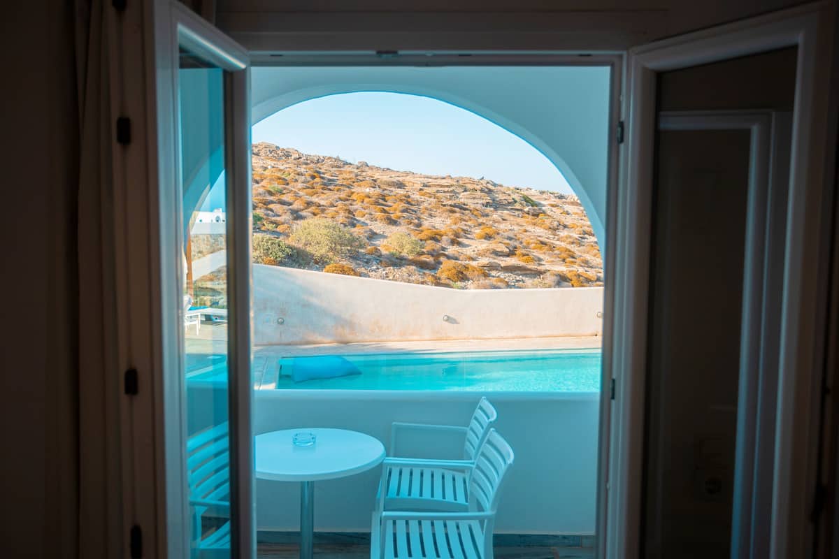 Vista a través de un arco de entrada hacia un área de piscina con una silla y mesa blancas, con vistas a una colina con vegetación escasa.
