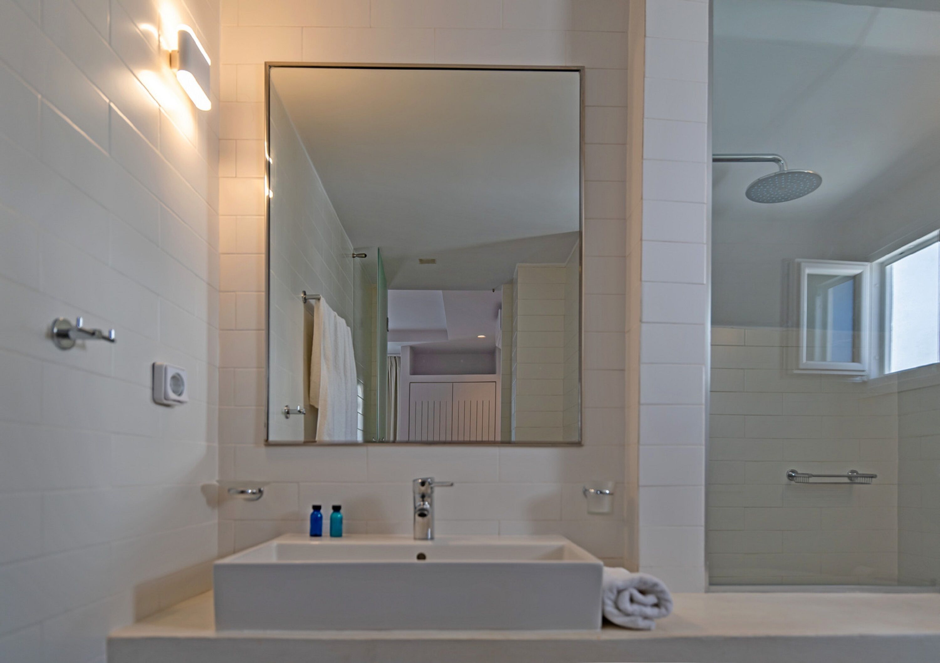 A modern bathroom with a large mirror, white tiles, a sink with two bottles, and a towel.