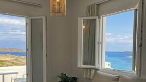 A room with a beige sofa, a hanging wicker light fixture, a potted plant, and a view of the sea through a window. - Adonis Hotel & Apartments (Paros)