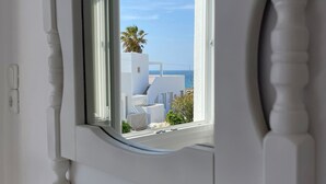 A window with a view of a white building, a palm tree, and the sea. - Adonis Hotel & Apartments (Paros)