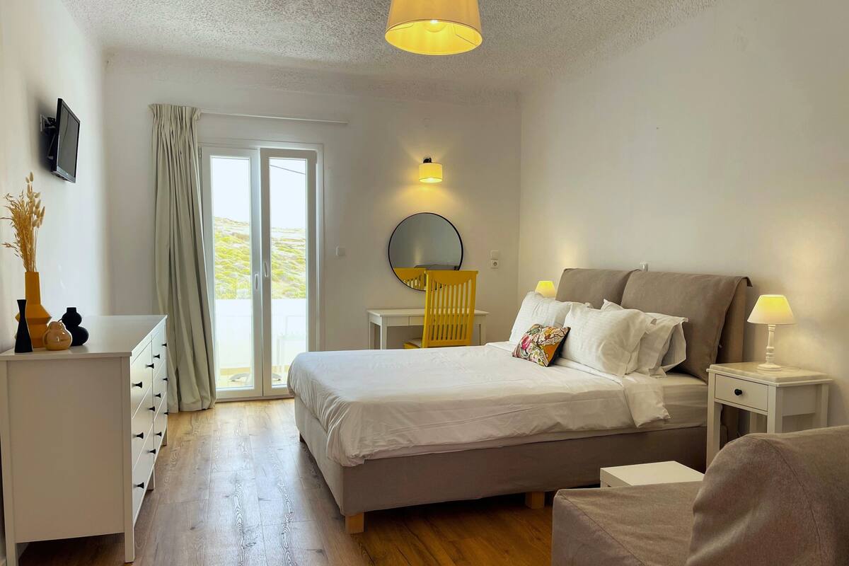 Habitación de hotel con cama, mesitas de noche, escritorio, silla, espejo y vistas al exterior.