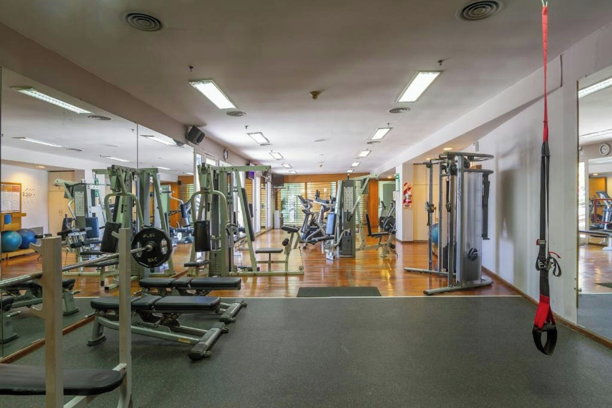 A gym with various exercise equipment, including weight machines and free weights, and a mirror wall.