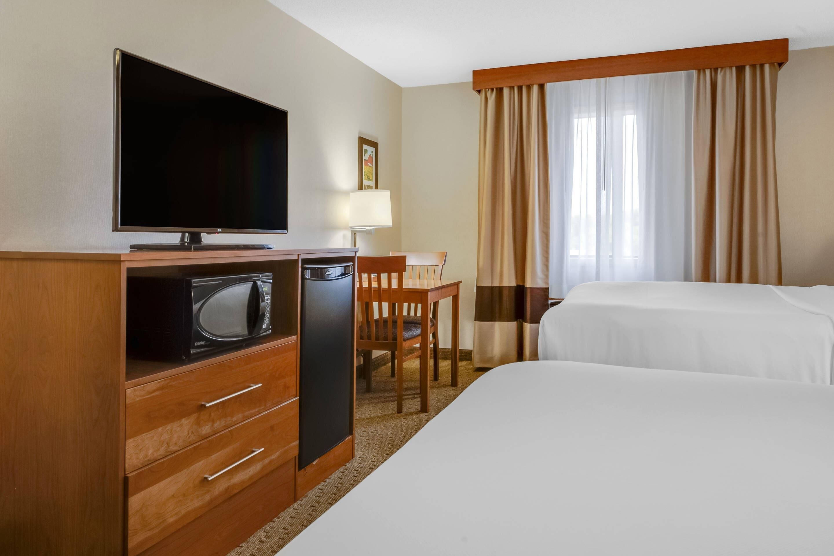 Quarto de hotel com televisão, micro-ondas e frigorífico na área de cozinha, uma mesa com cadeira e duas camas.