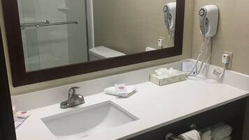Baño con un espejo grande, lavamanos, toallero, secador de pelo y un cubo de basura.
