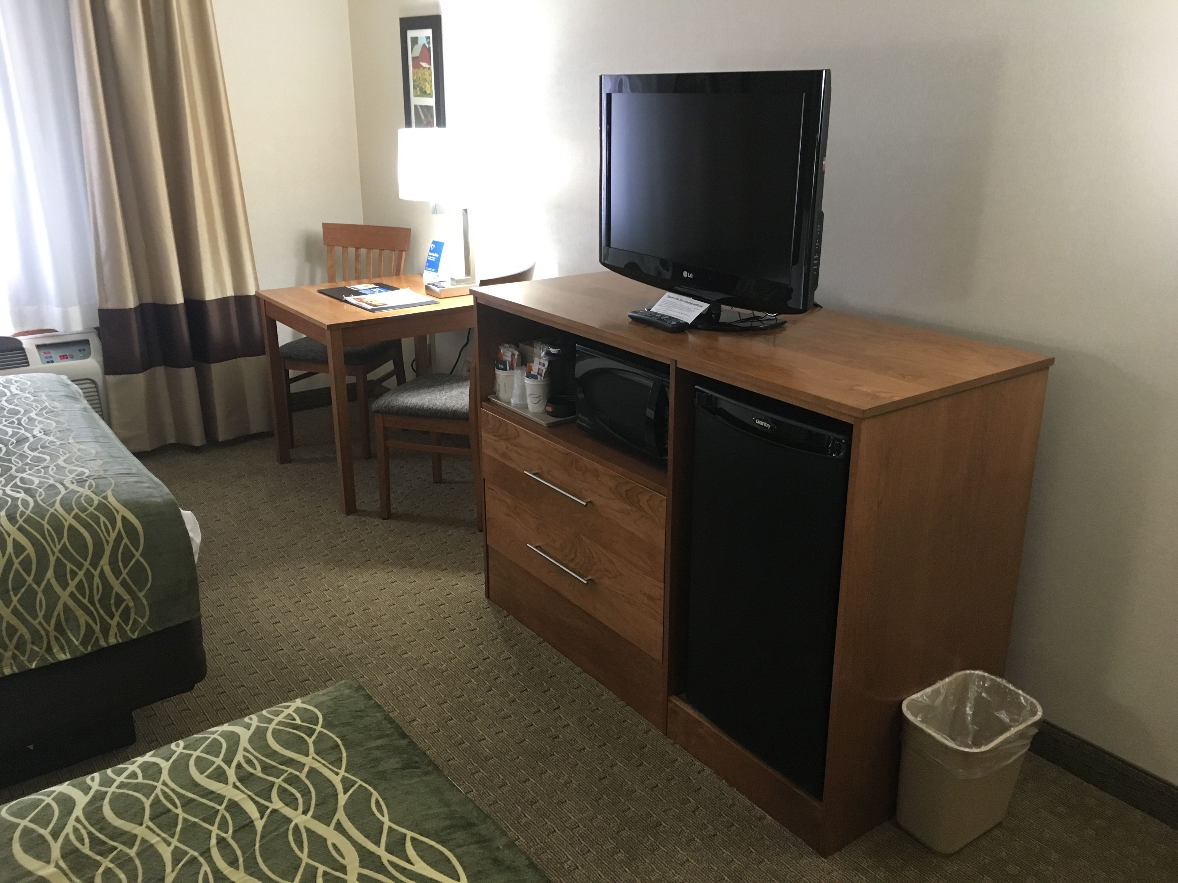 Habitación de hotel con un escritorio de madera, televisor, refrigerador pequeño, una silla, cama y un cesto de basura.