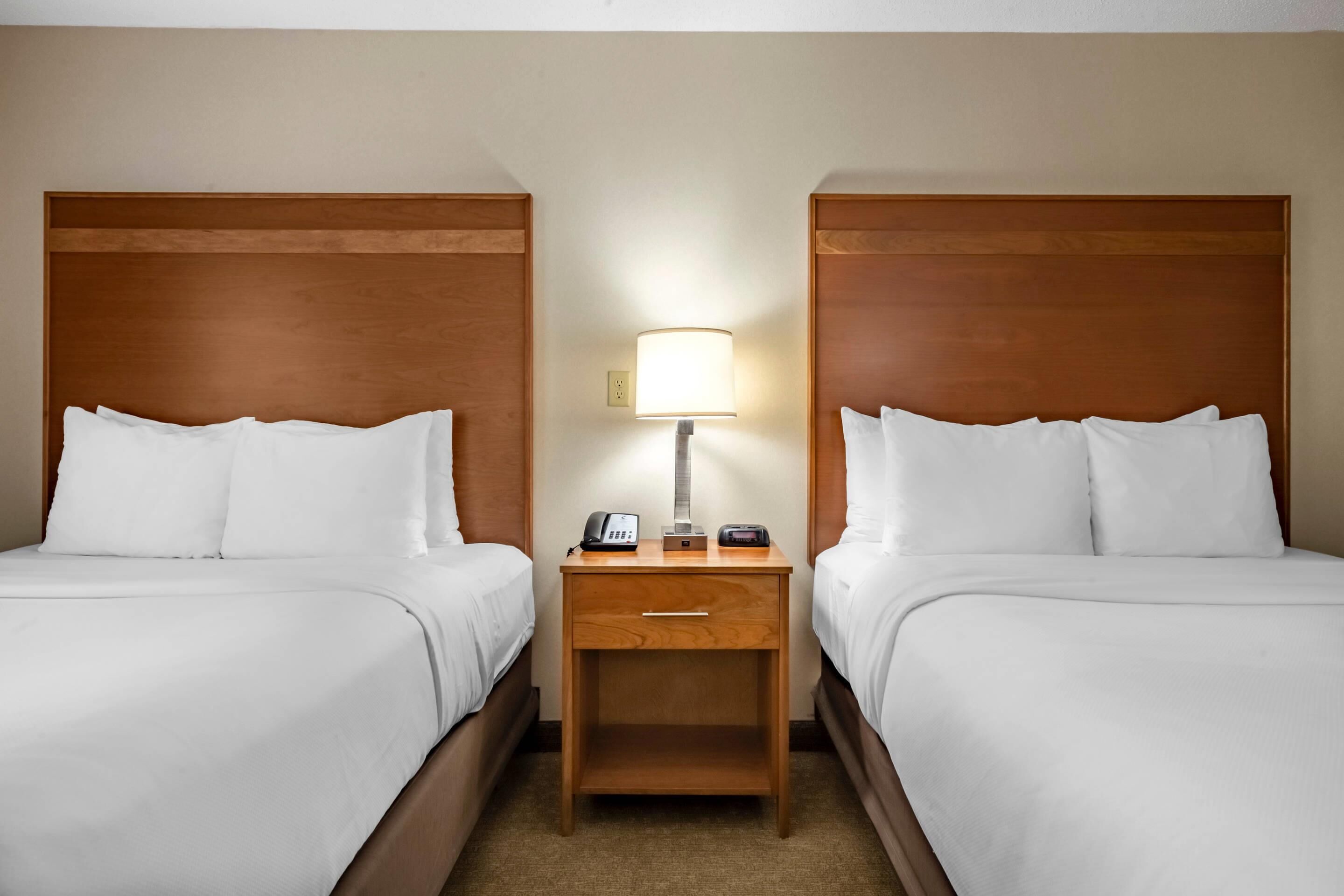 A hotel room with two beds, a nightstand with a lamp, a phone, and a digital clock.