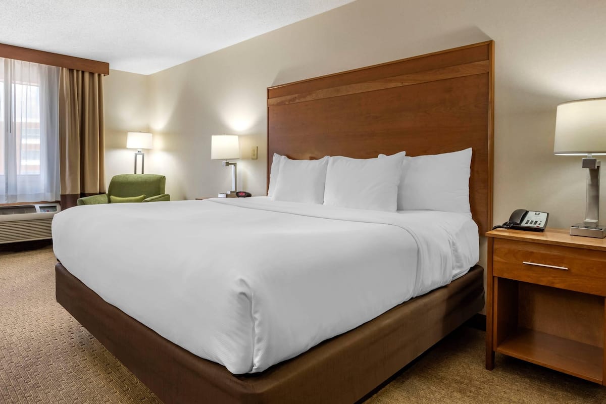 A hotel room with a large bed, a wooden headboard, two bedside tables with lamps, a green chair, and a window with curtains.