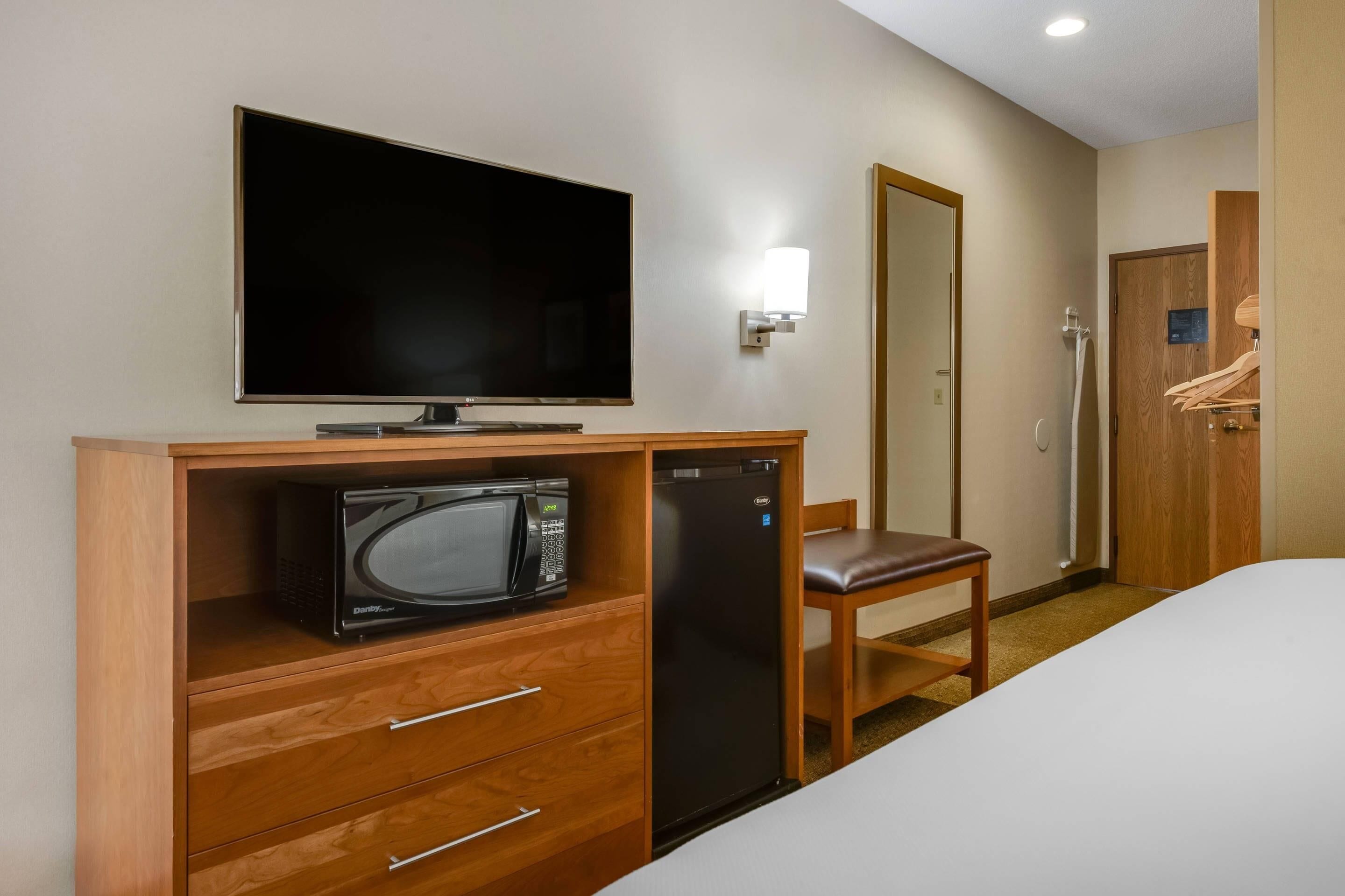 Habitación de hotel con televisión de pantalla plana, microondas, refrigerador, escritorio de madera, silla y armario.